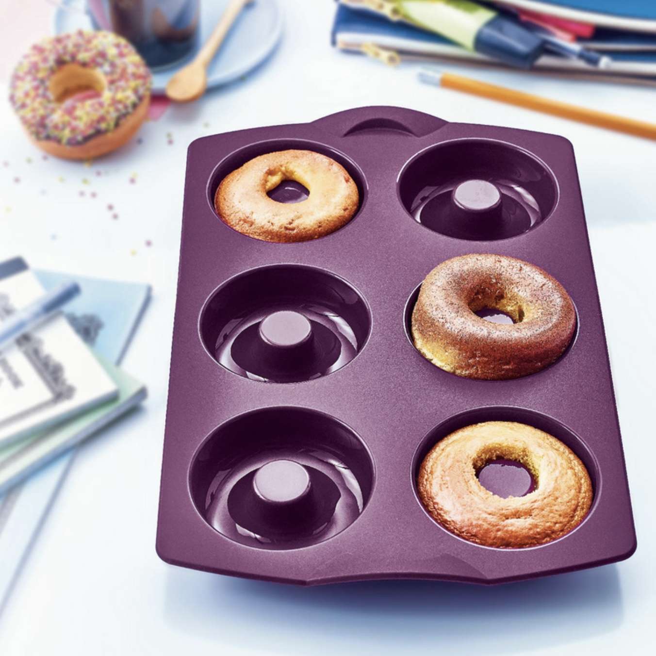 Purple silicone donut baking mold with three baked donuts, kitchen tools in background