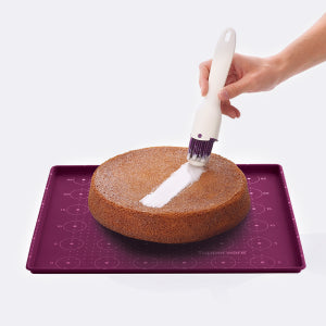 Hand using a silicone pastry brush to apply glaze on a round sponge cake on a purple baking mat