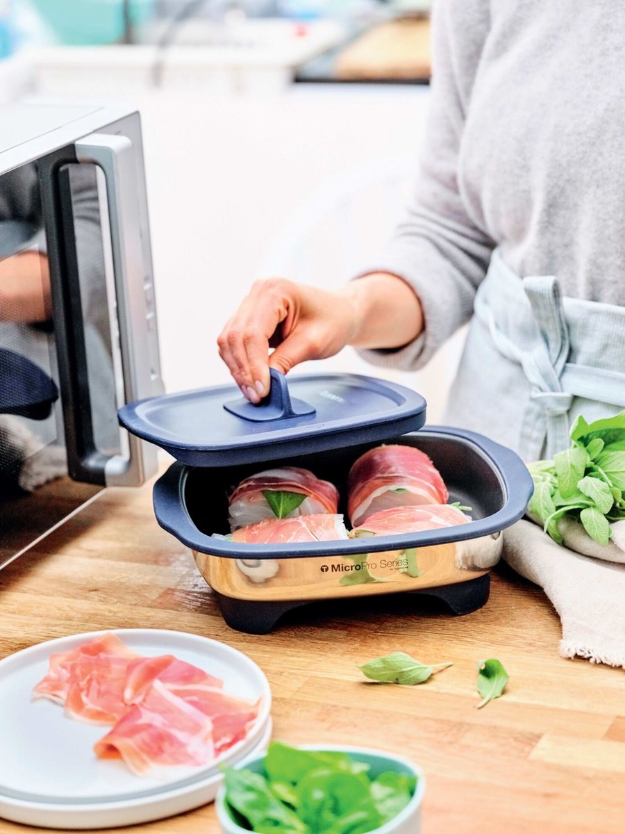 Person using a microwave-safe cooking dish with prosciutto-wrapped food and fresh herbs