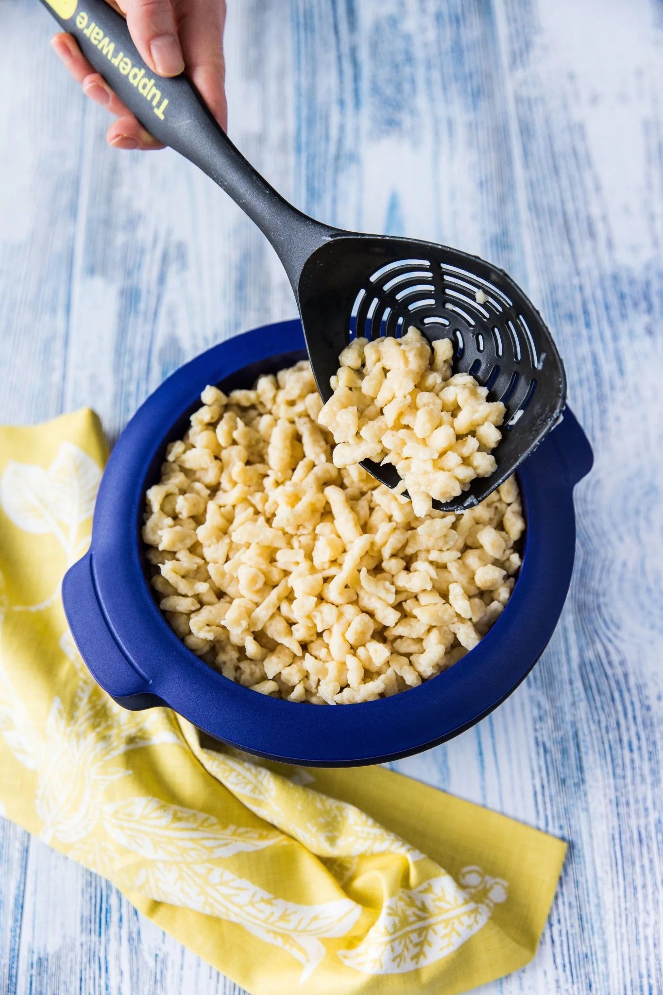 Slotted spoon lifting pasta from blue bowl on wooden table with yellow cloth