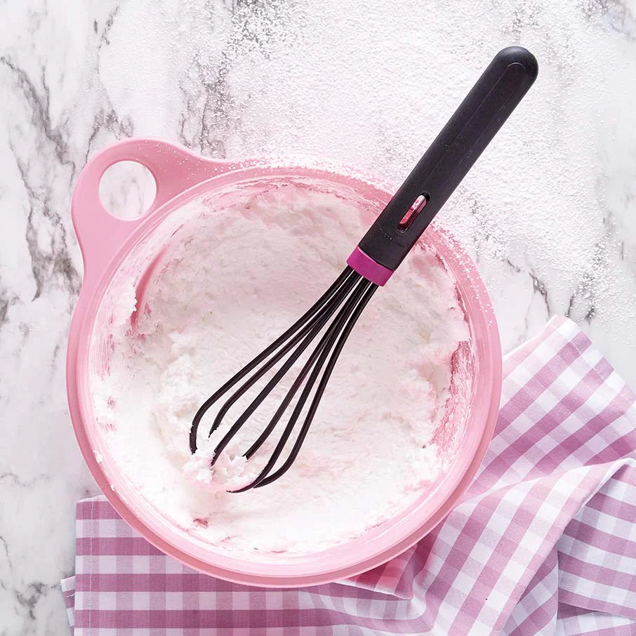 Black whisk in pink mixing bowl with whipped cream on checkered kitchen towel