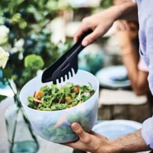 Handen serveren verse salade met zwarte keukentongs in een witte kom