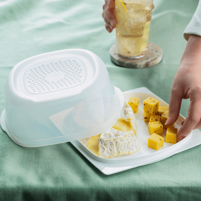 Rectangular cheese storage container with assorted cheeses on a white tray, hand reaching for cheese cubes