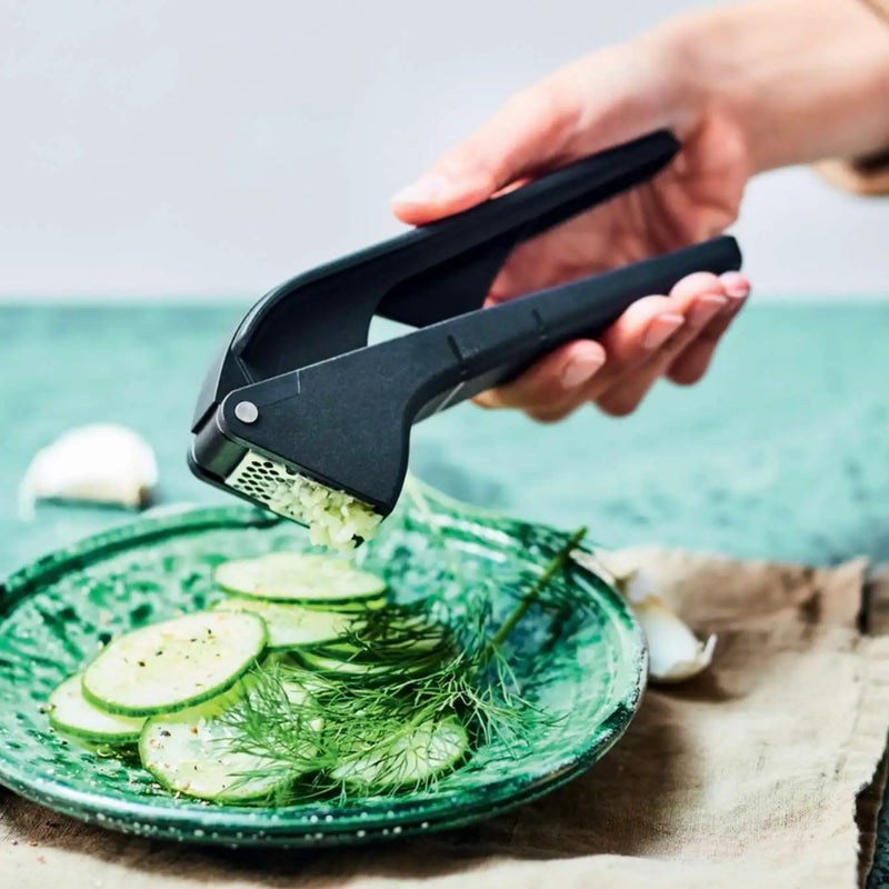 Hand using a black Tupperware garlic press to mince garlic over sliced cucumbers on a green plate.