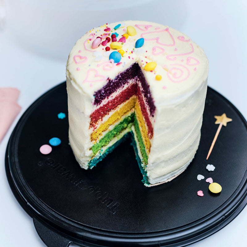 Rainbow layered cake with white frosting on a black Tupperware platter.