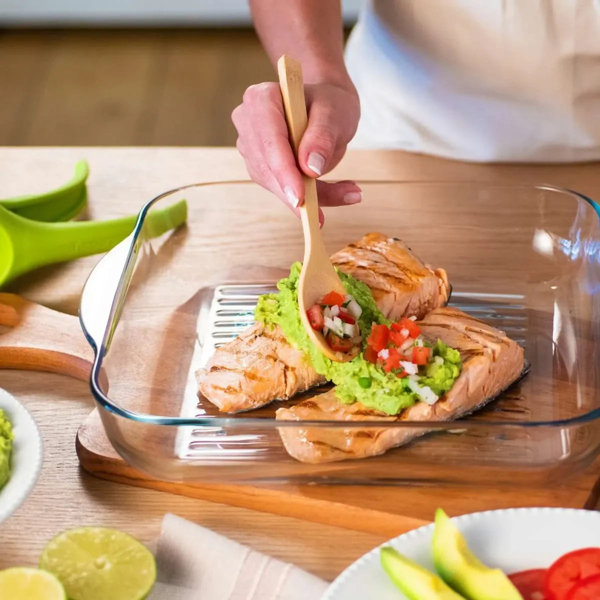 Grilled salmon fillets topped with guacamole and salsa in glass baking dish, wooden spoon in use