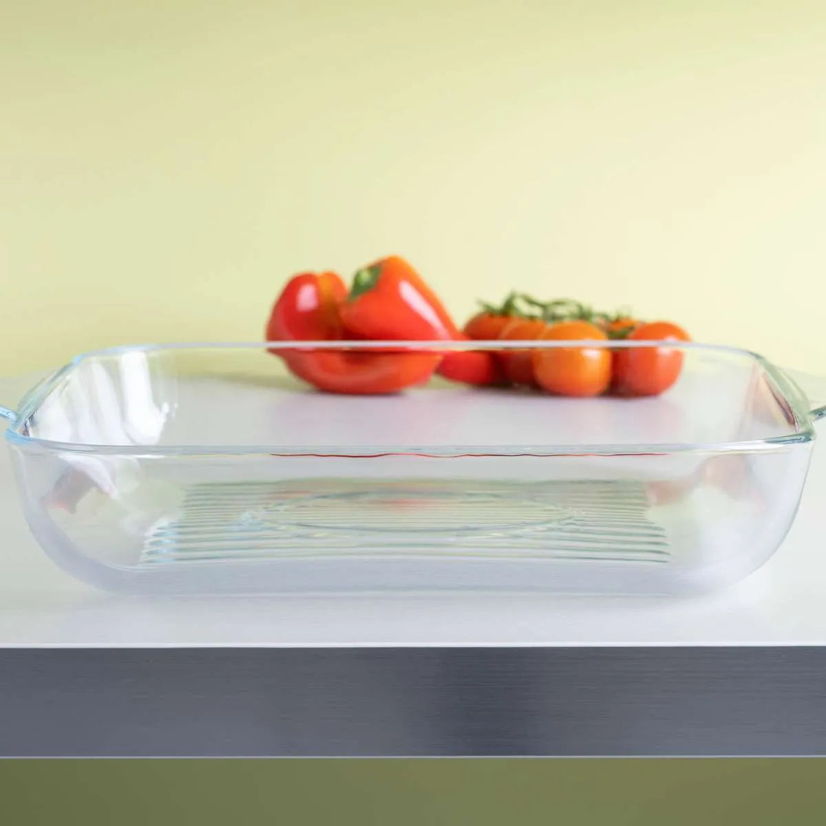 Clear glass baking dish on kitchen counter with red bell peppers and tomatoes in background