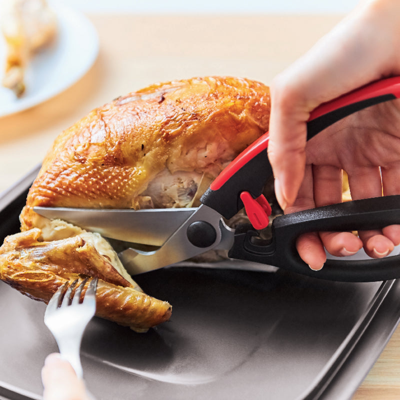 A person using Tupperware poultry shears to carve a roasted chicken on a dark tray.