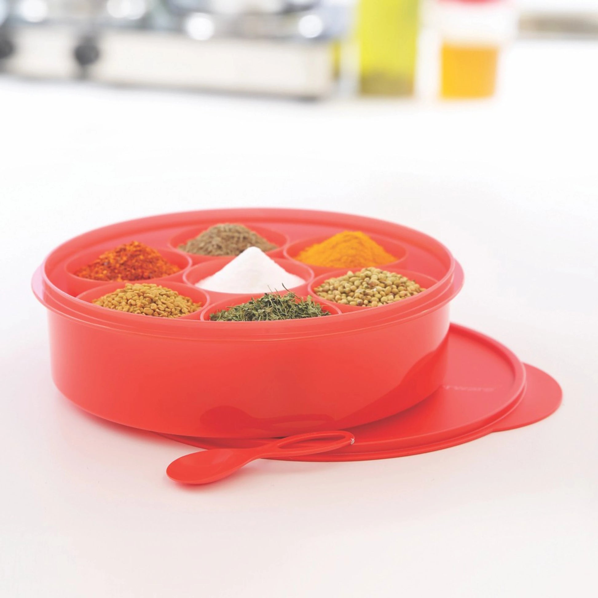Red plastic spice box with multiple compartments filled with assorted spices and a matching spoon