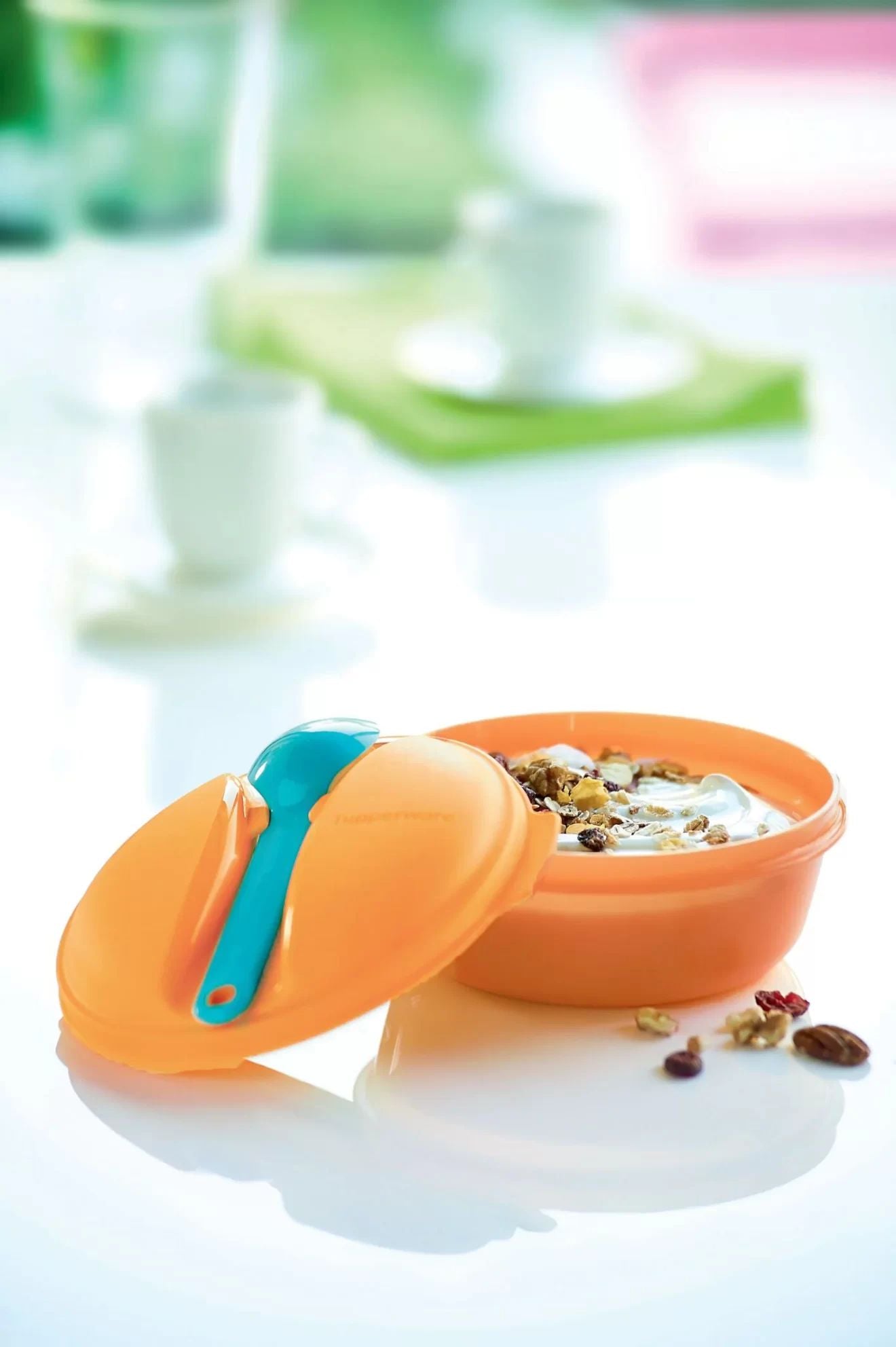 Orange plastic food container with blue spoon, filled with yogurt and granola, on a white table.