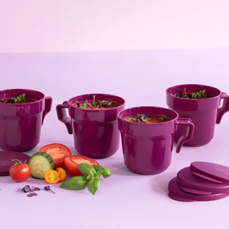 Set of four purple soup mugs with handles and lids, fresh vegetables in foreground on a light surface