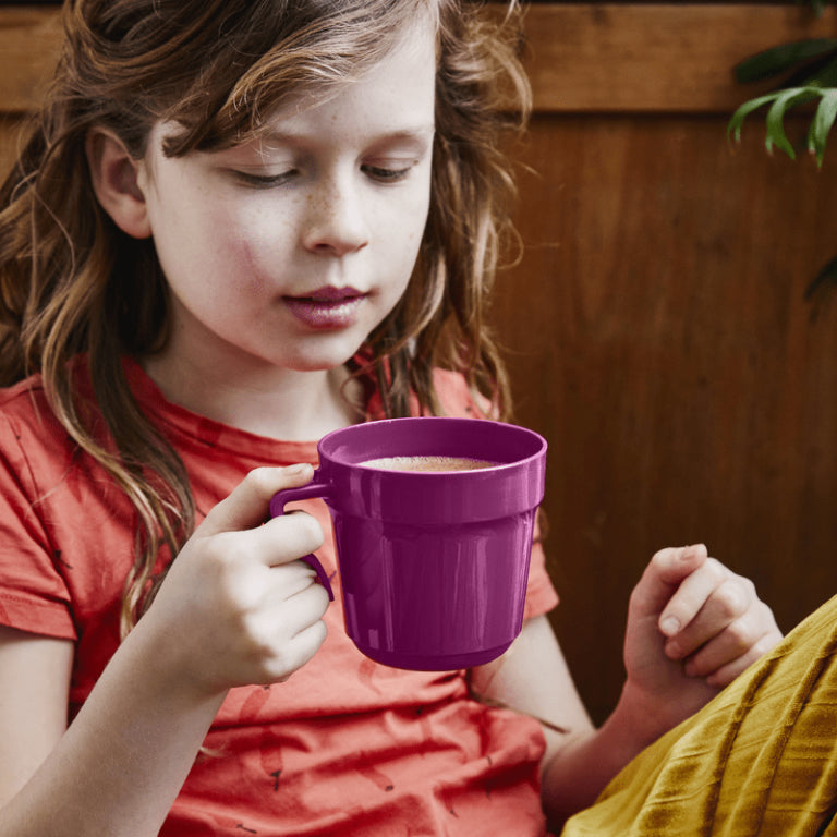 Child holding a purple plastic mug with a warm drink, seated indoors