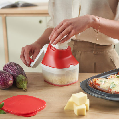 Person using red and white manual cheese grater in kitchen with vegetables and lasagna nearby