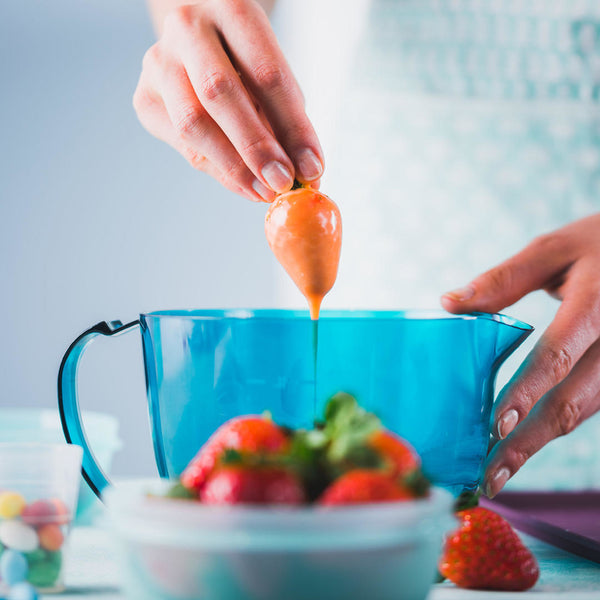 Dipping a fresh strawberry in melted chocolate using a blue mixing bowl in a kitchen setting