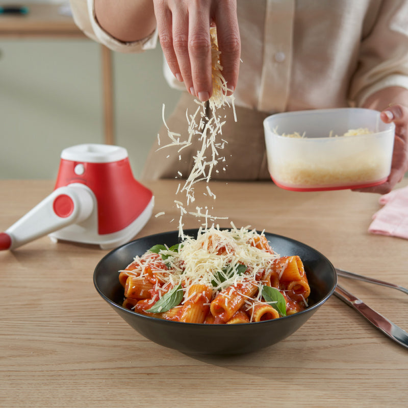 Hand sprinkling fresh grated cheese over pasta in a black bowl with cheese grater tool nearby