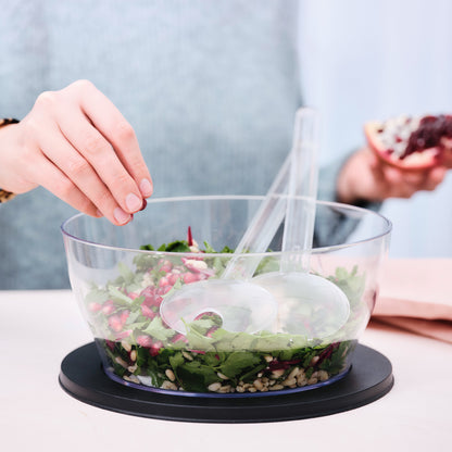 Hand adding pomegranate seeds to fresh salad in a clear mixing bowl with salad servers