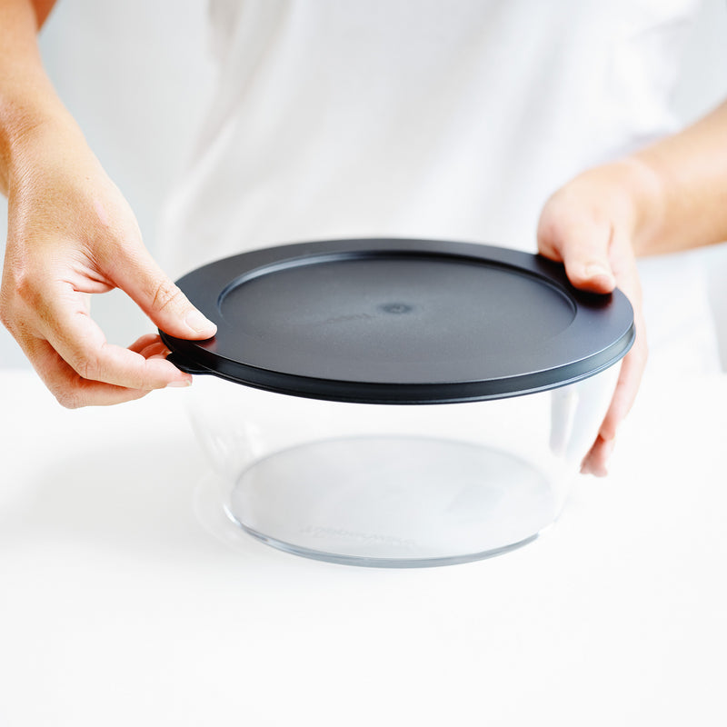 Hands closing a round clear food storage container with a black lid on a white surface