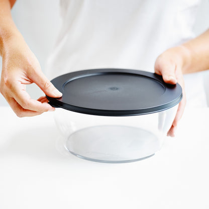 Hands closing a round clear food storage container with a black lid on a white surface