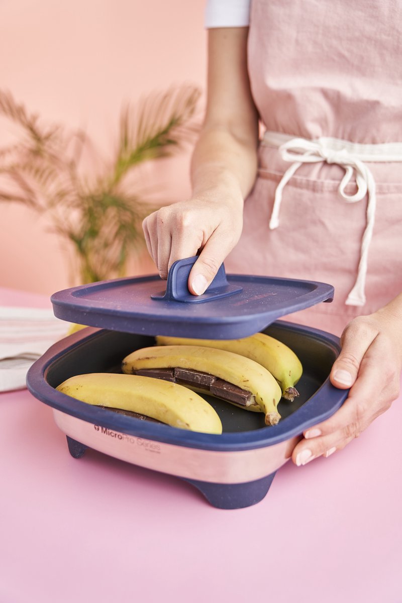 Person opening a microwave-safe dish with bananas and chocolate bars inside, on a pink background