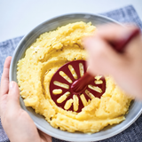 Rode Tupperware aardappelstamper gebruikt om aardappelen te pureren in een grijze kom.