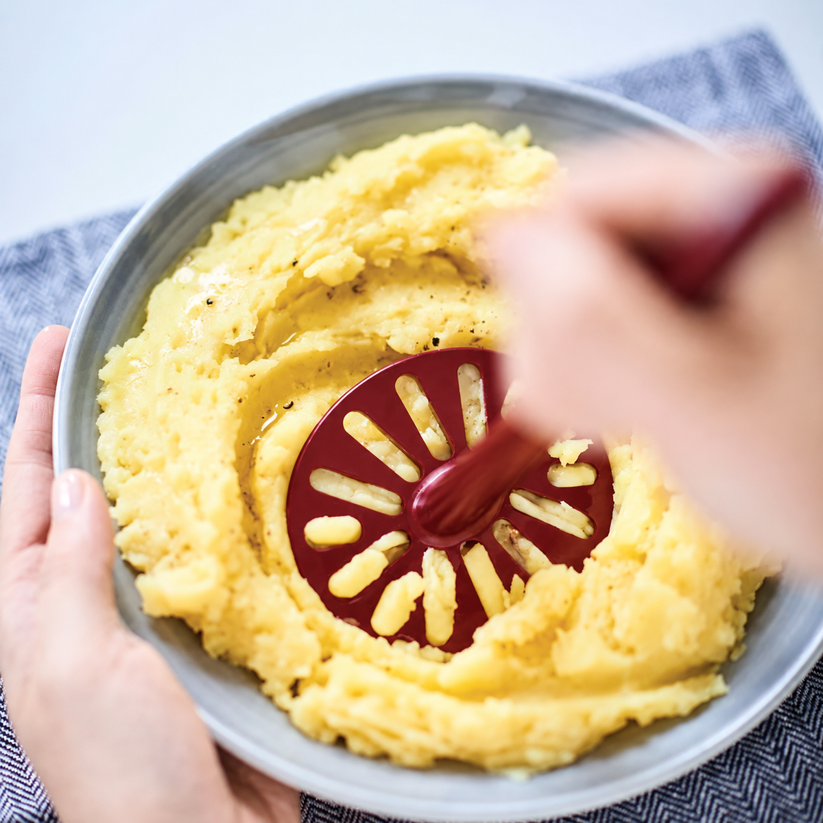 Rode Tupperware aardappelstamper gebruikt om aardappelen te pureren in een grijze kom.