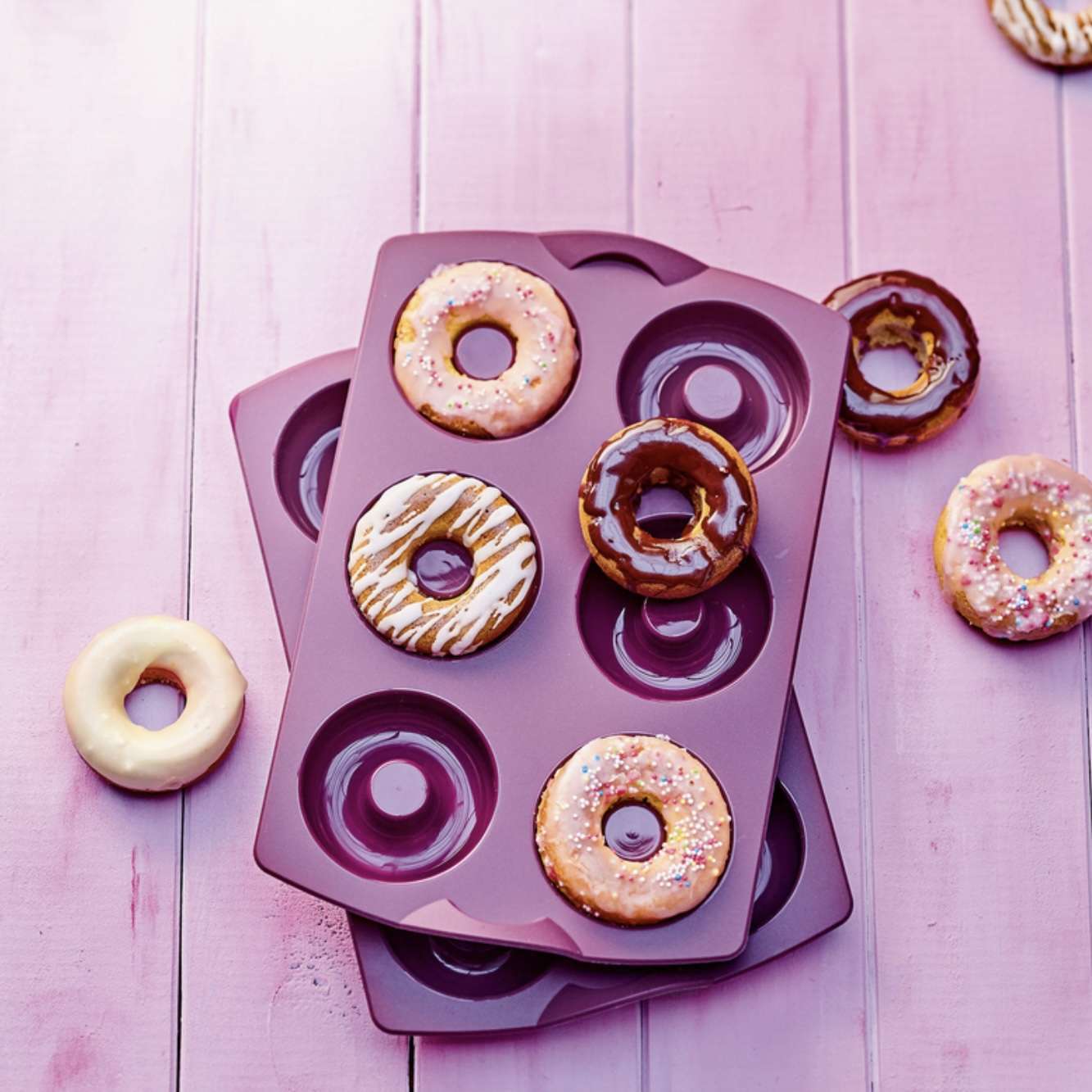 Purple silicone donut baking molds with decorated donuts on pink wooden surface