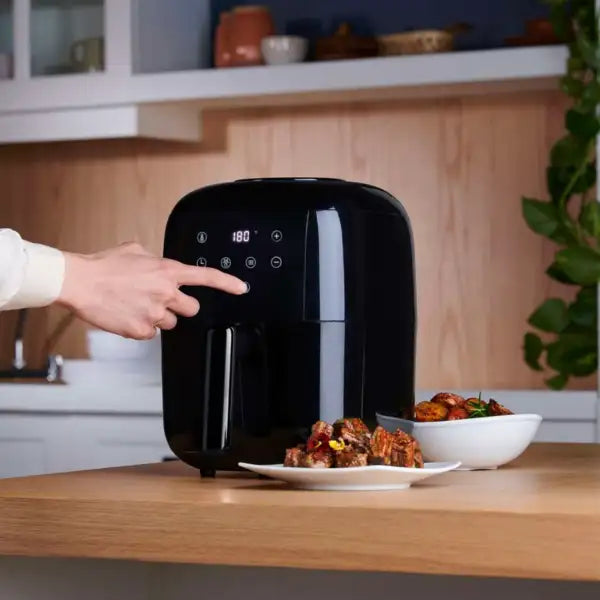 Modern black air fryer with digital display in kitchen, person selecting setting, plates of food nearby