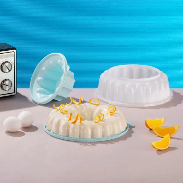 Bundt cakevormen in lichtblauw en wit, met versierde cake, eieren en sinaasappelschijfjes op keukentafel