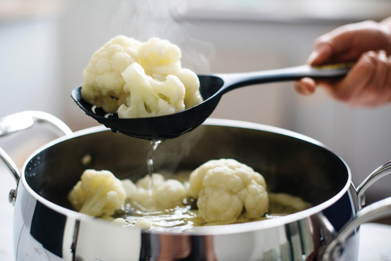 Black slotted spoon lifting steamed cauliflower from a stainless steel pot in a kitchen.