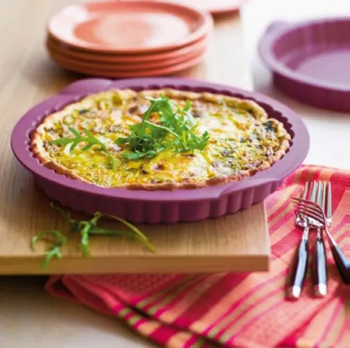 Fresh quiche in purple silicone baking pan on wooden board with plates and cutlery