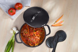 Cast iron casserole pot with tomato stew and vegetables on a light wooden table