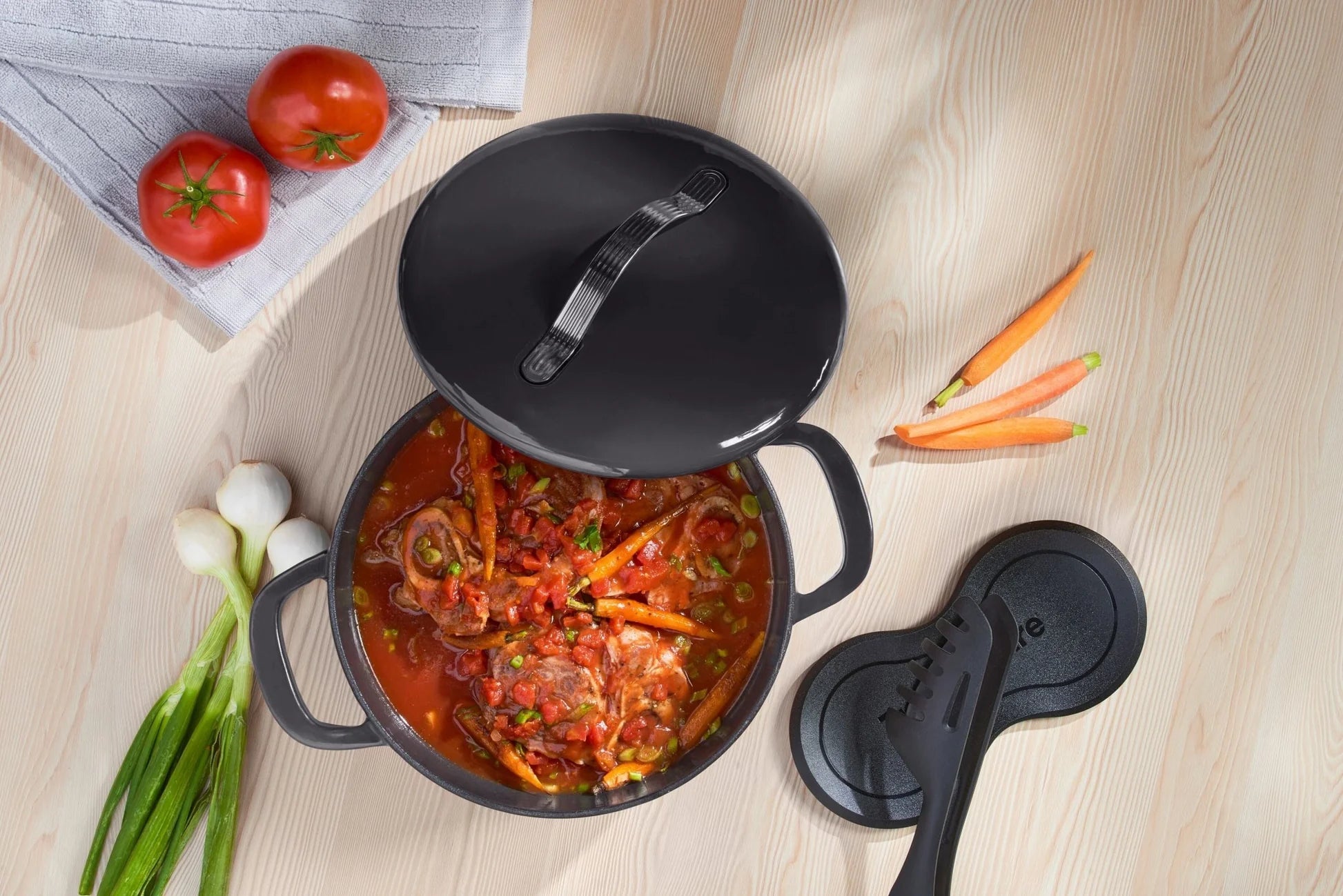 Cast iron casserole pot with tomato stew and vegetables on a light wooden table