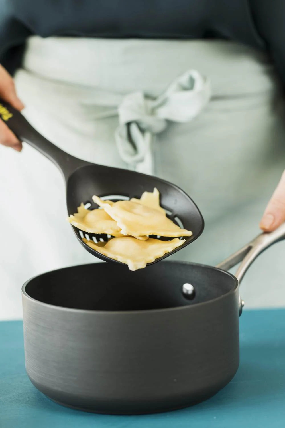 Black slotted spoon lifting cooked ravioli from a saucepan in a kitchen setting