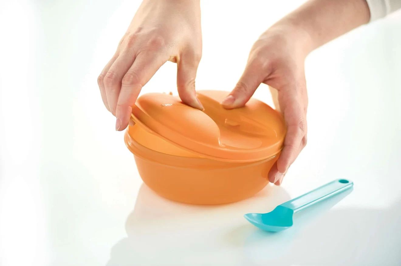 Hands closing an orange microwave-safe food container with lid, blue kitchen spoon nearby