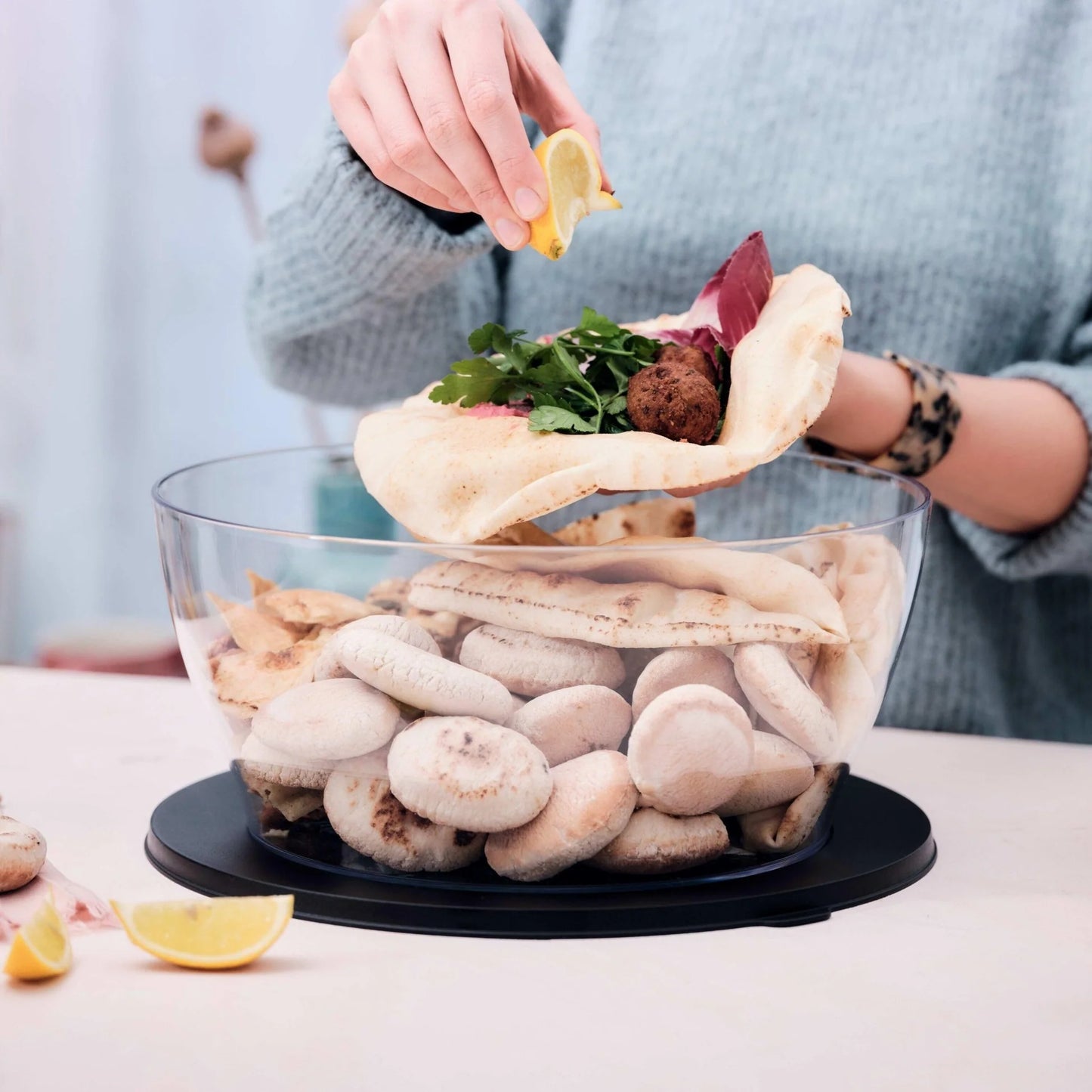 Clear plastic salad bowl filled with pita bread, person squeezing lemon over stuffed pita