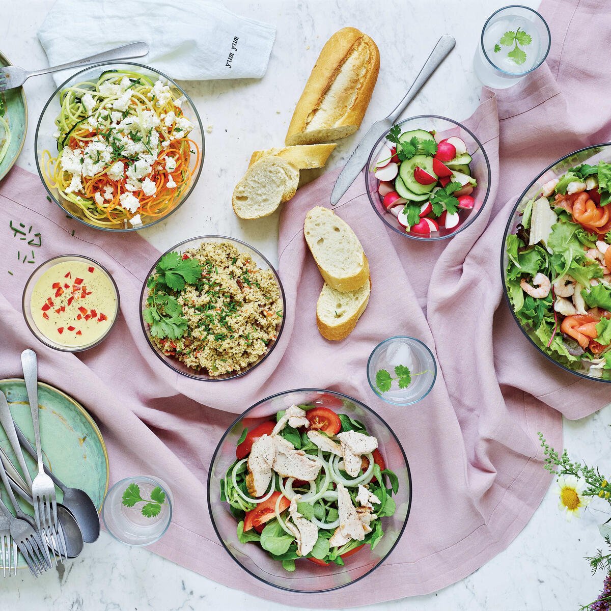 Clear eco-friendly salad bowls with fresh salads, bread, and water on a marble table.