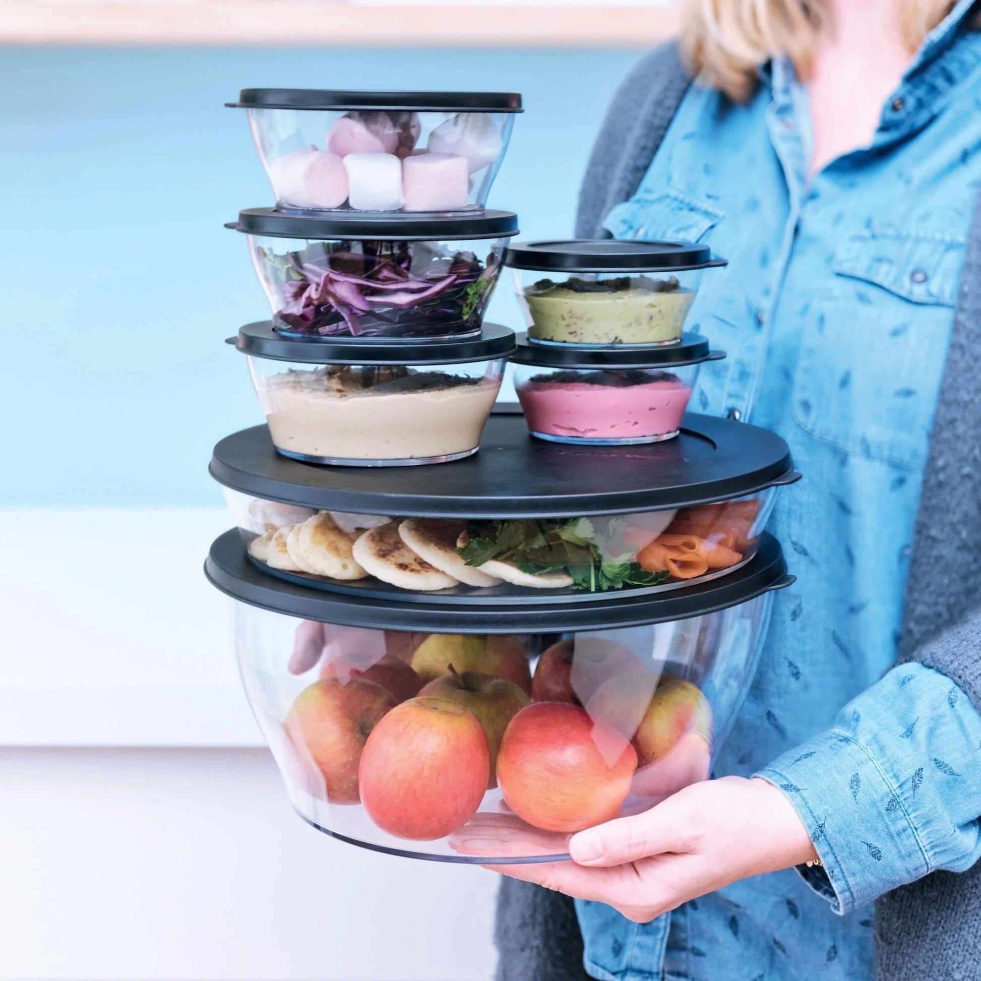 Stack of clear eco-friendly salad bowls with lids, filled with fruits and snacks, held by a person