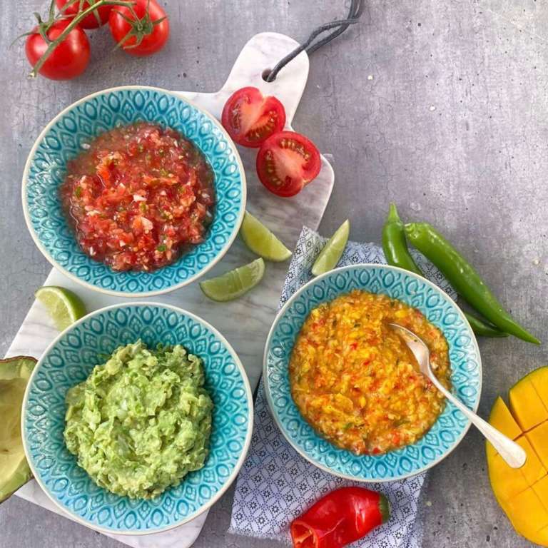 Three colorful dips—guacamole, tomato salsa, mango salsa—in patterned bowls with fresh lime, tomato, chili, and mango on a kitchen countertop.