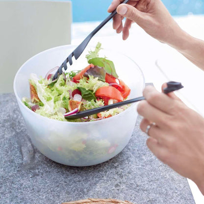 Zwarte salade tongs gebruikt om verse groenten te mengen in een transparante kom