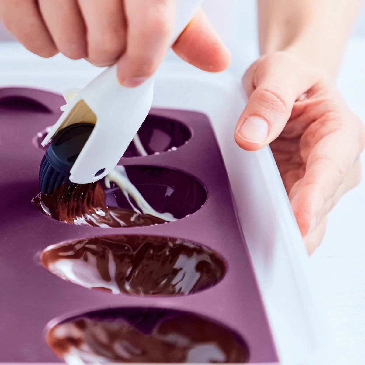 Hand brushing melted chocolate in a purple silicone egg mold kitchen tool
