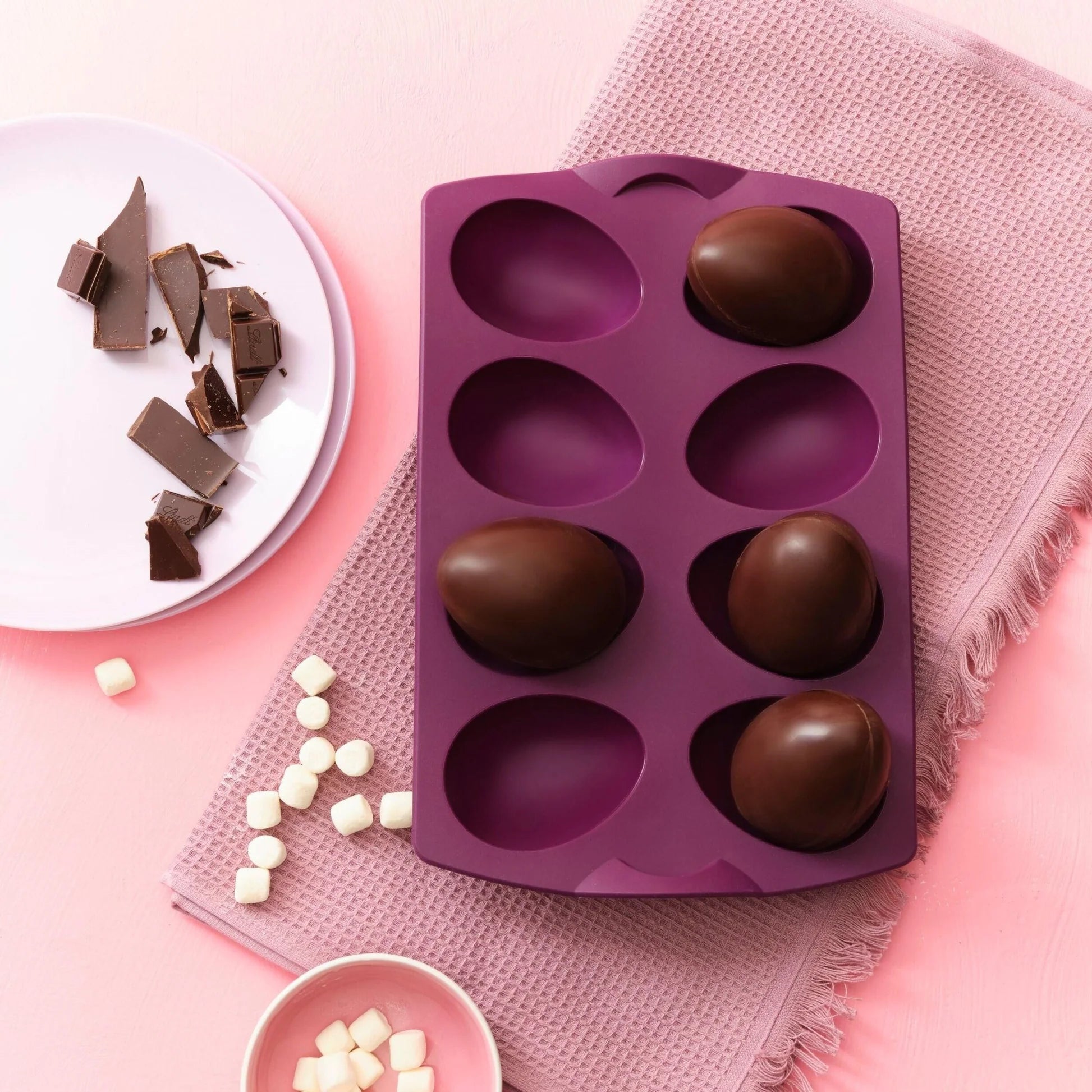 Purple silicone egg mold with chocolate eggs, plate of chocolate pieces, and marshmallows on pink background
