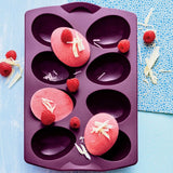 Purple silicone egg-shaped baking mold with pink desserts, raspberries, and white chocolate shavings