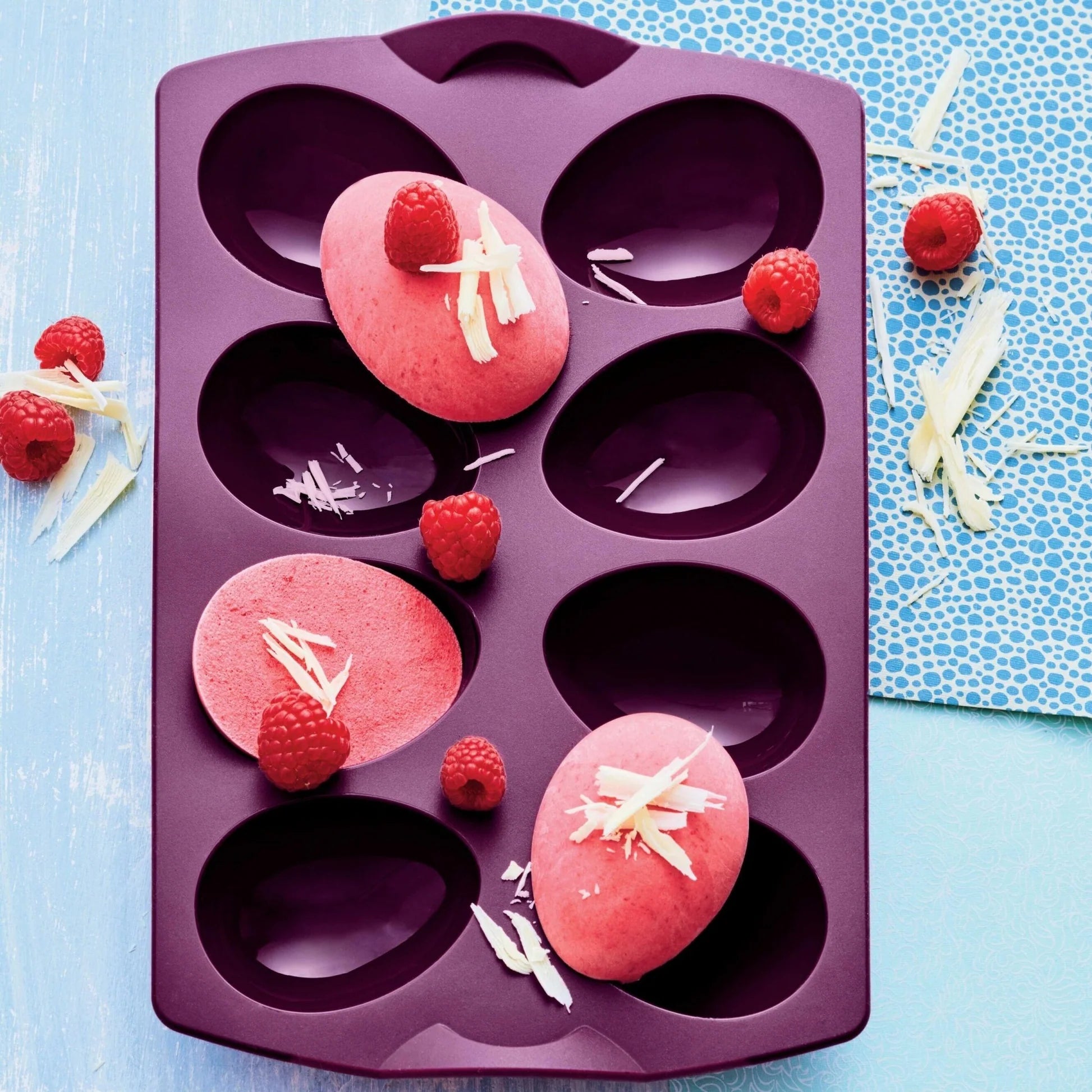 Purple silicone egg-shaped baking mold with pink desserts, raspberries, and white chocolate shavings