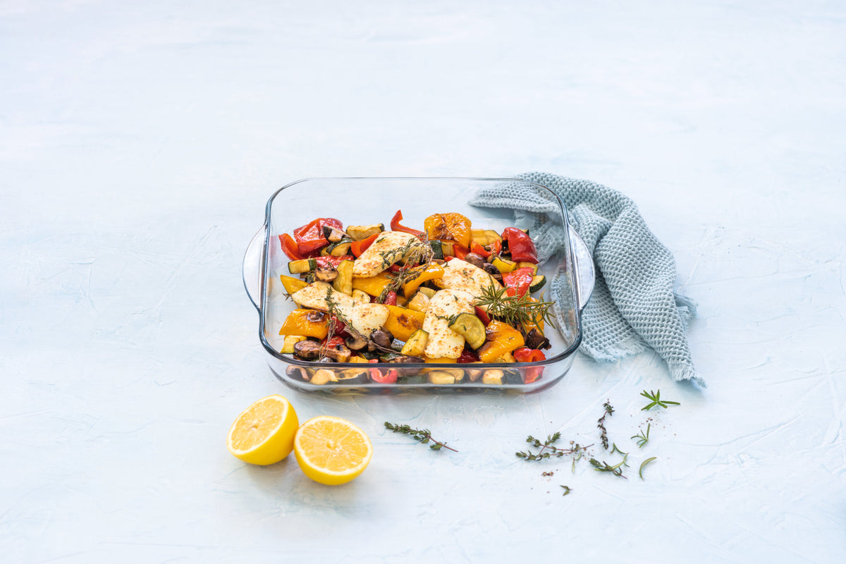 Glass baking dish with roasted vegetables, herbs and lemon on light blue background