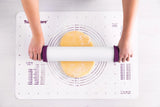 Hands using a non-stick rolling pin with purple handles to roll dough on a pastry mat