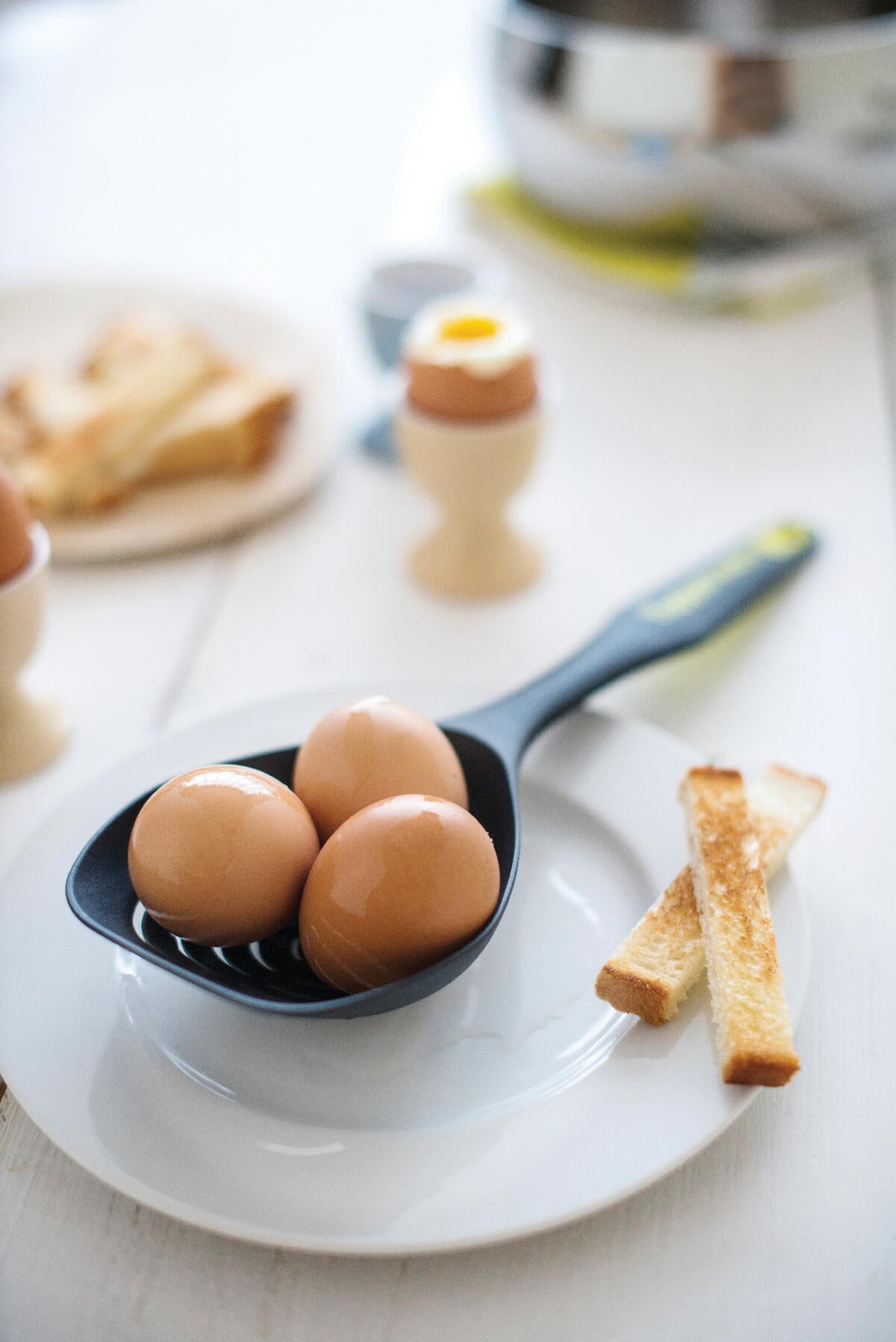 Black skimmer spoon with three boiled eggs on white plate, next to toast soldiers, breakfast setting