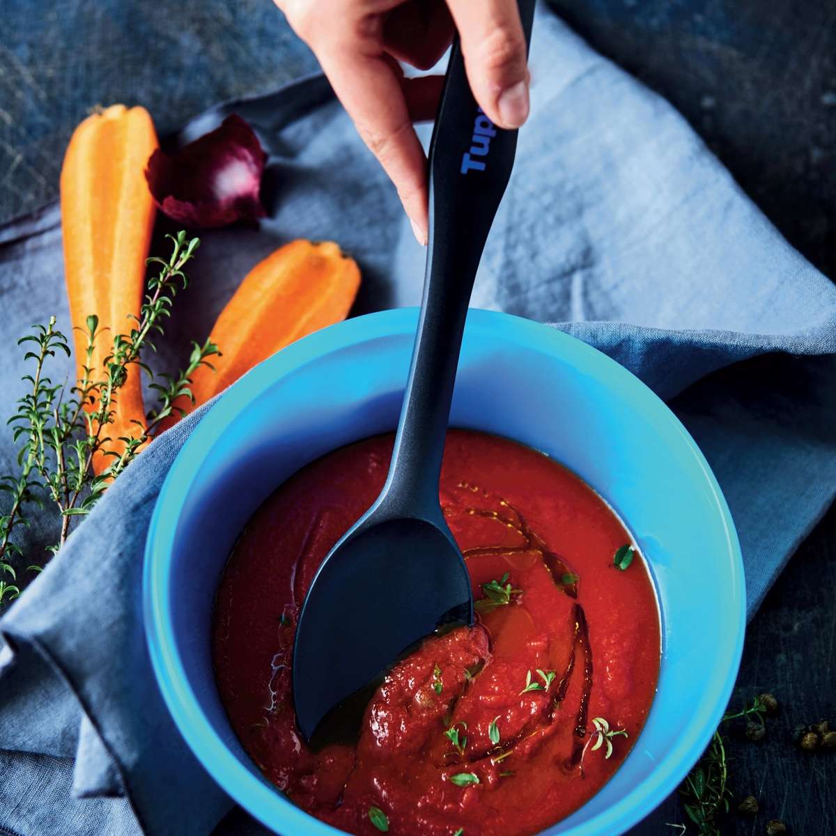 Black kitchen spoon stirring tomato soup in blue bowl, carrots and herbs nearby