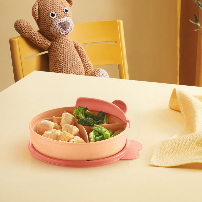 Roze kindermaaltijdplaat met vakken voor groenten en kip, op tafel met knuffelbeer