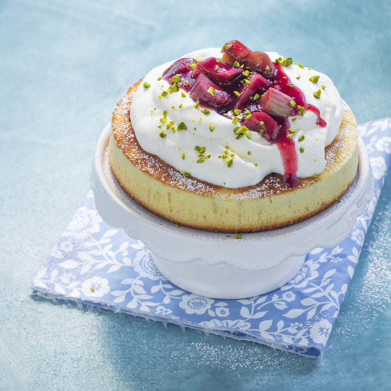 A small cake with whipped cream, rhubarb topping, and pistachios on a white Tupperware stand.