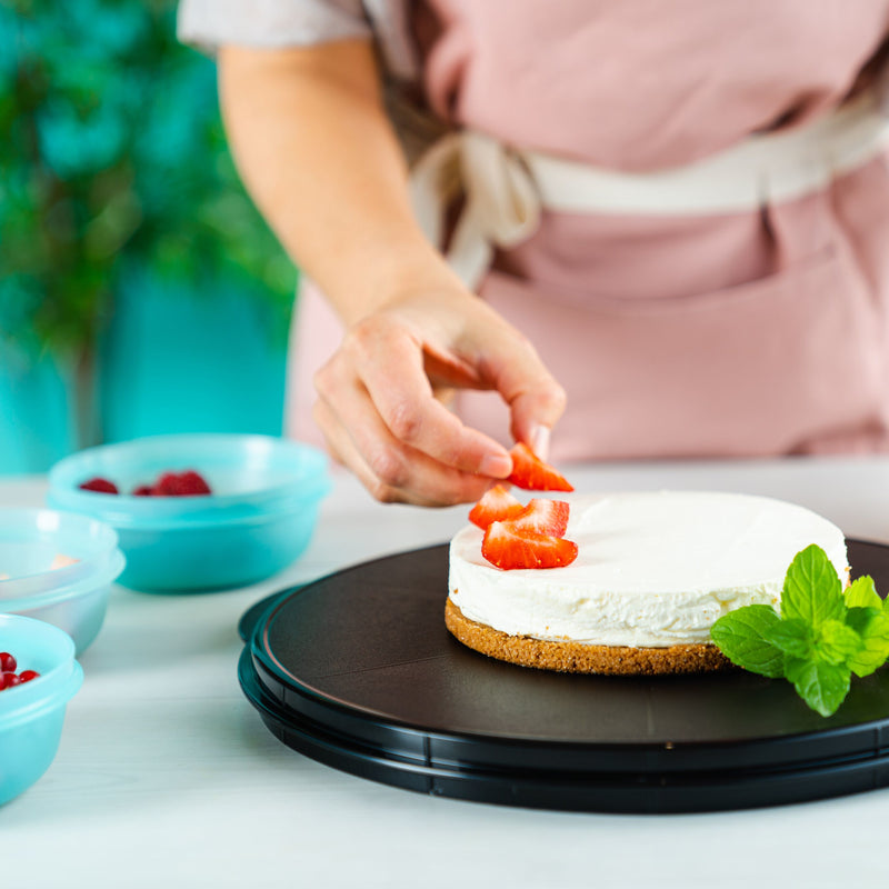 A person decorating a cheesecake with sliced strawberries on a black platter.